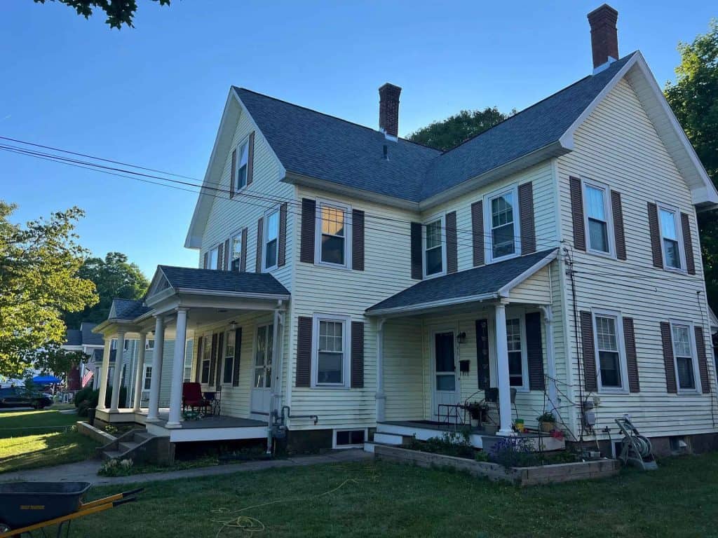 A large, two-story yellow house with a steeply pitched roof and brown shutters. The house has multiple chimneys and front porches. There's a bicycle and a yellow wheelbarrow on the grass in the well-maintained yard. Trees and clear blue sky are visible in the background, showcasing expert gutter installation by York County specialists.