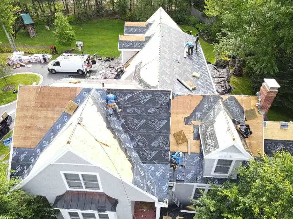 Aerial view of workers installing a new roof on a white house surrounded by trees. Roofing materials and tools are scattered, with parts covered in black synthetic underlayment. A white work van and additional supplies, likely for siding replacement in York County, are visible on the driveway.